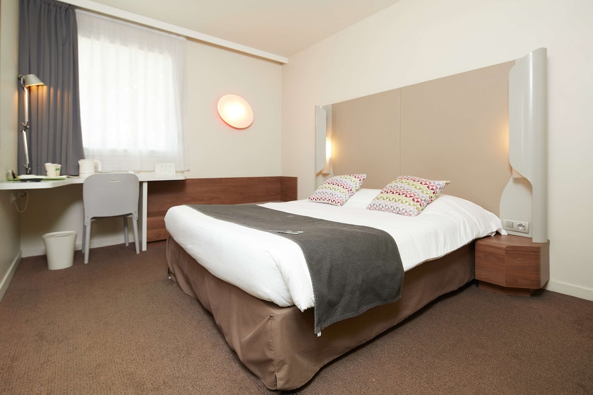 A hotel room with a bed, bedside table, lamp, desk, chair, and a window with curtains.