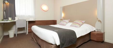 A hotel room with a bed, bedside table, lamp, desk, chair, and a window with curtains.