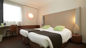 Habitación de hotel con dos camas, un escritorio, una silla, un cesto de basura y una mesita de noche con lámpara.