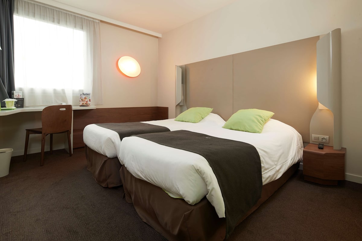 A hotel room with two beds, a desk, a chair, a trash bin, and a bedside table with a lamp.