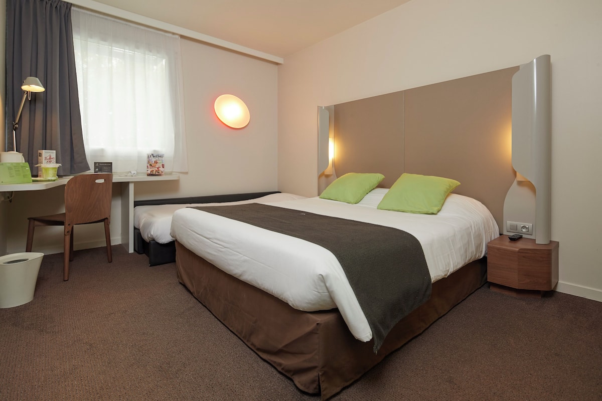 A hotel room with a bed, desk, chair, and a small table with a lamp.