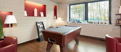 A pool table in a room with wooden flooring, red chairs, and a window with a view of trees.