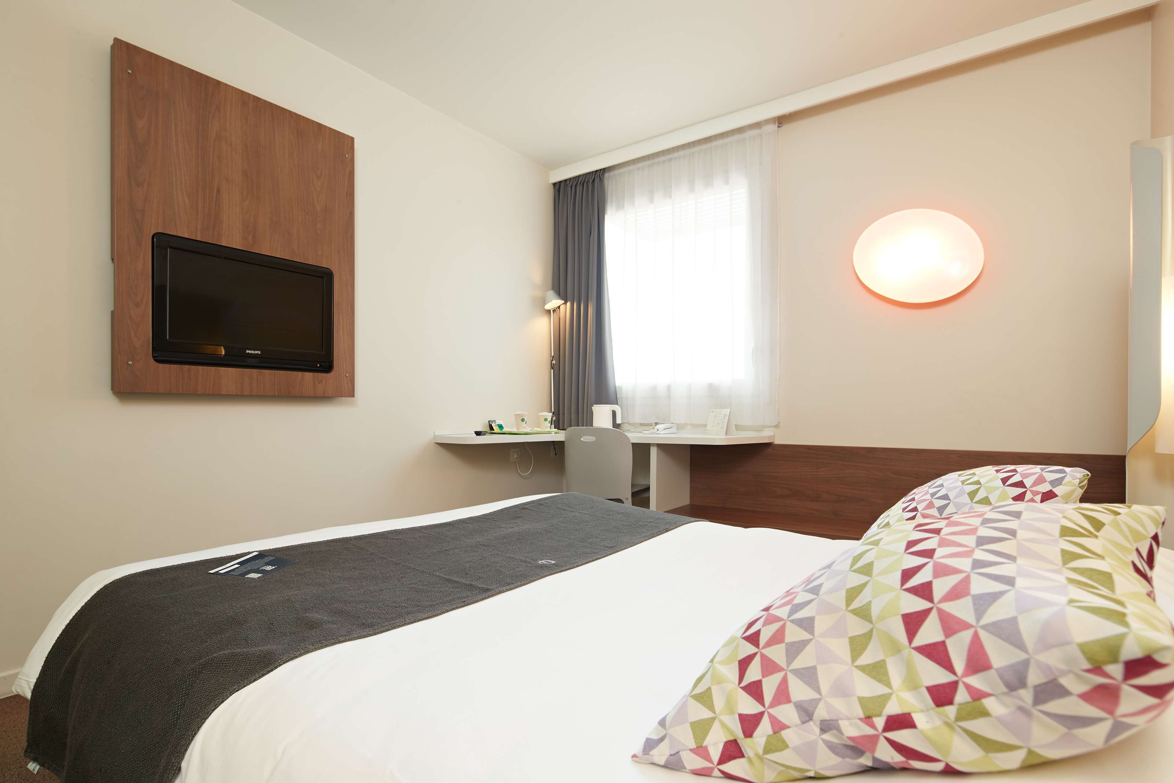 Une chambre d’hôtel comprenant un lit, un bureau, un téléviseur à écran plat, une lampe de chevet et une fenêtre avec des rideaux.