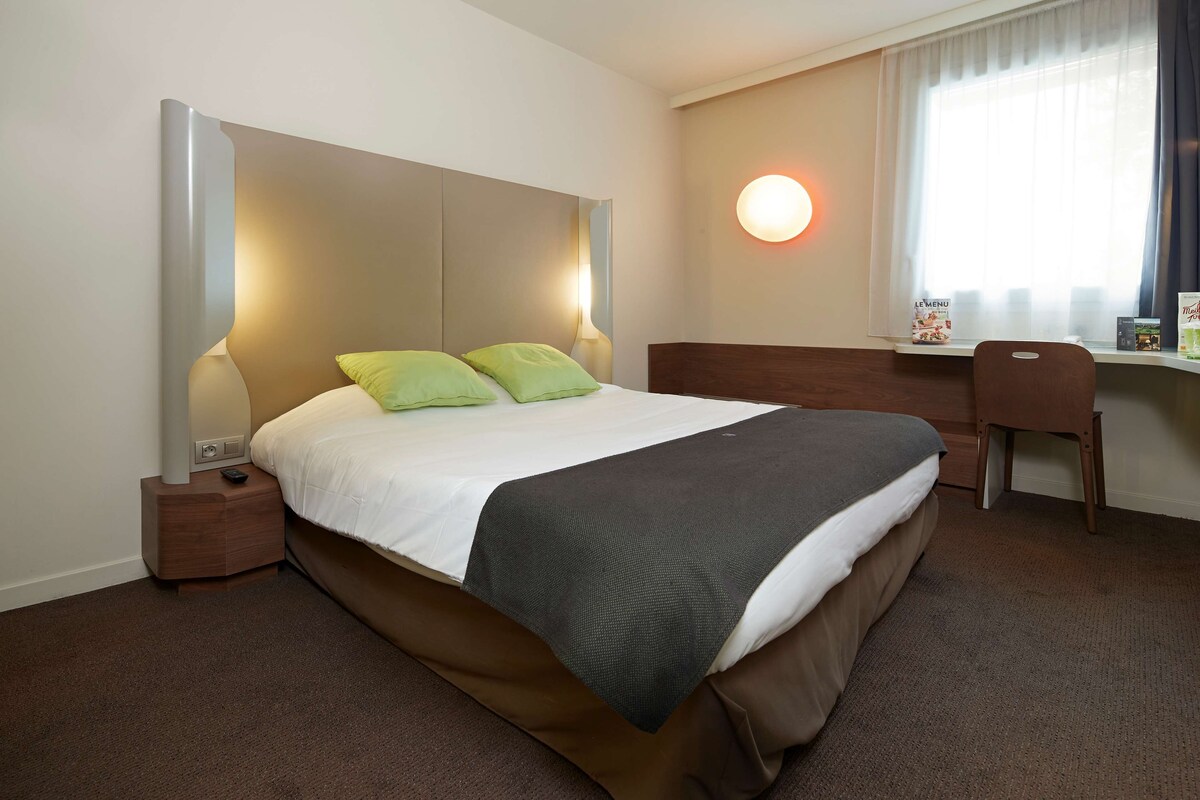 A hotel room with a large bed, a bedside table, a chair, a desk, and a window with curtains.