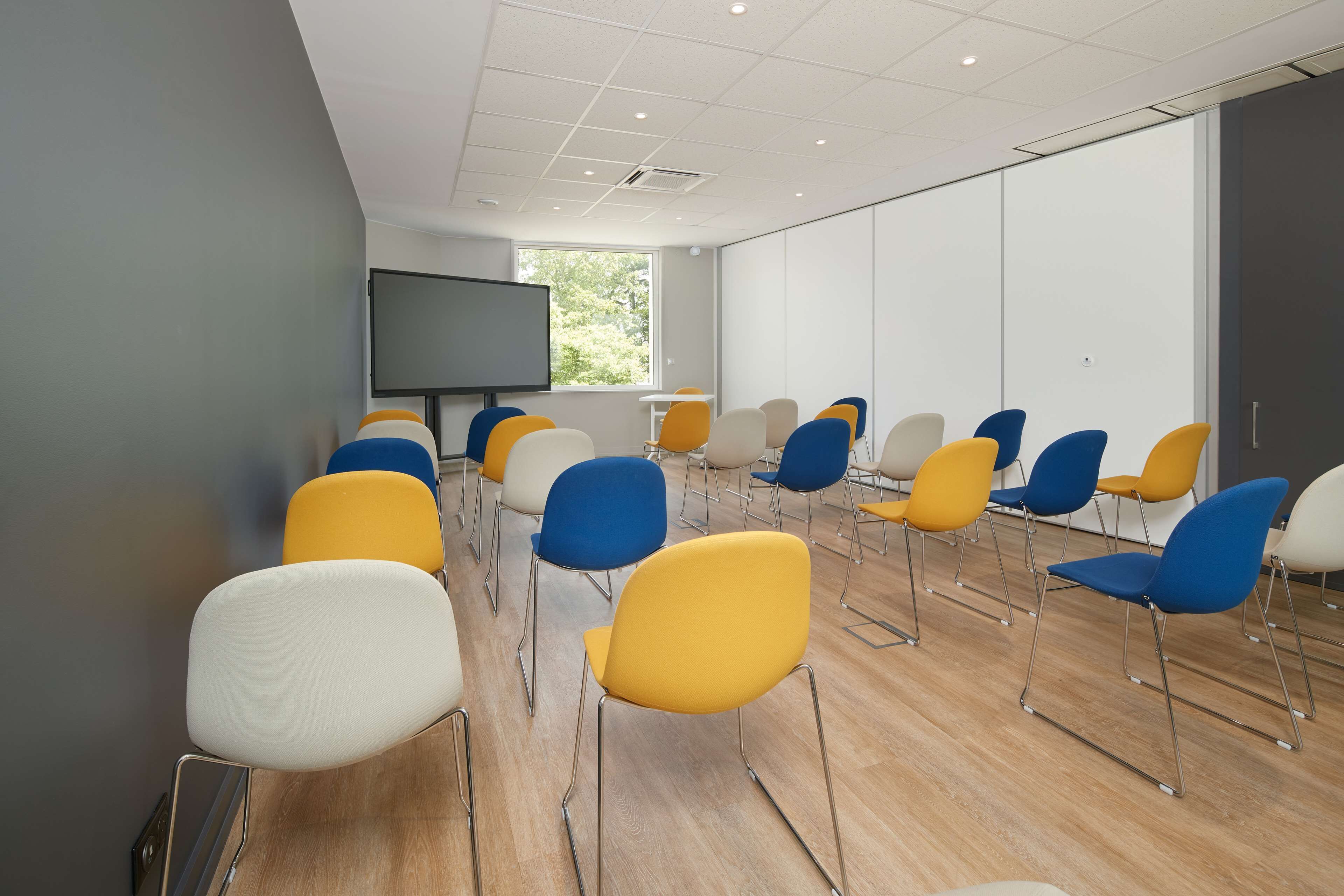 A conference room with rows of chairs, a large screen, and a window with a view of greenery.