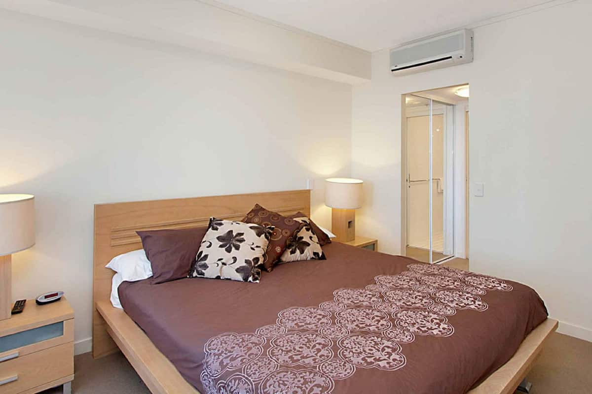 A bedroom with a bed, two bedside lamps, a nightstand, an air conditioning unit, and a door leading to a bathroom.
