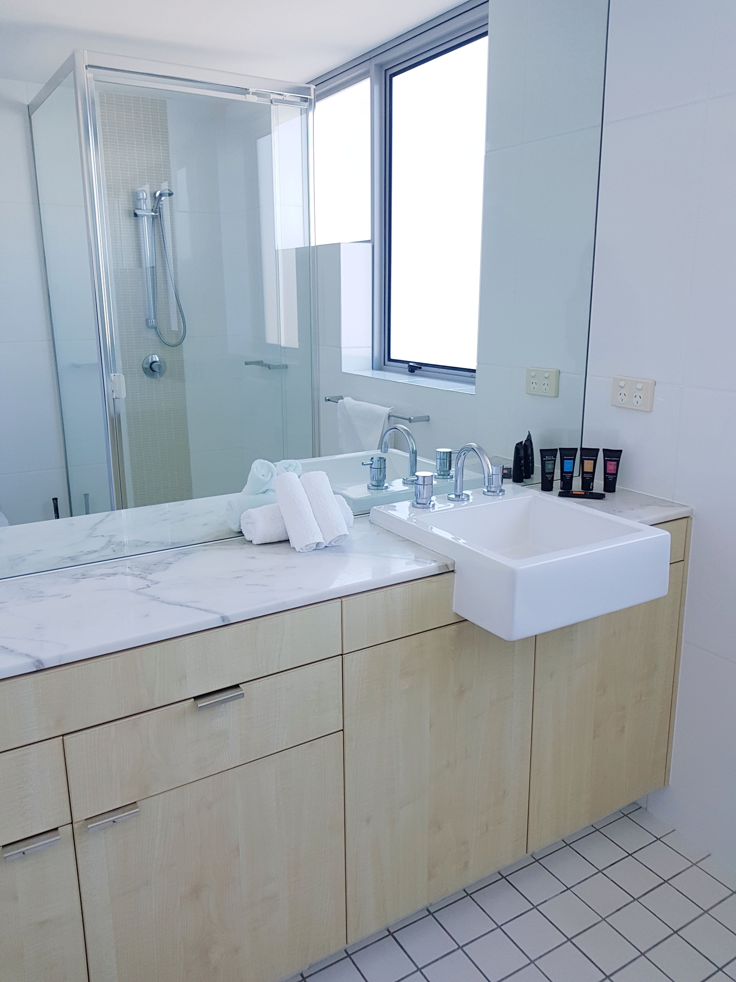 A modern bathroom with a large mirror, a glass shower enclosure, a white sink, and a window.