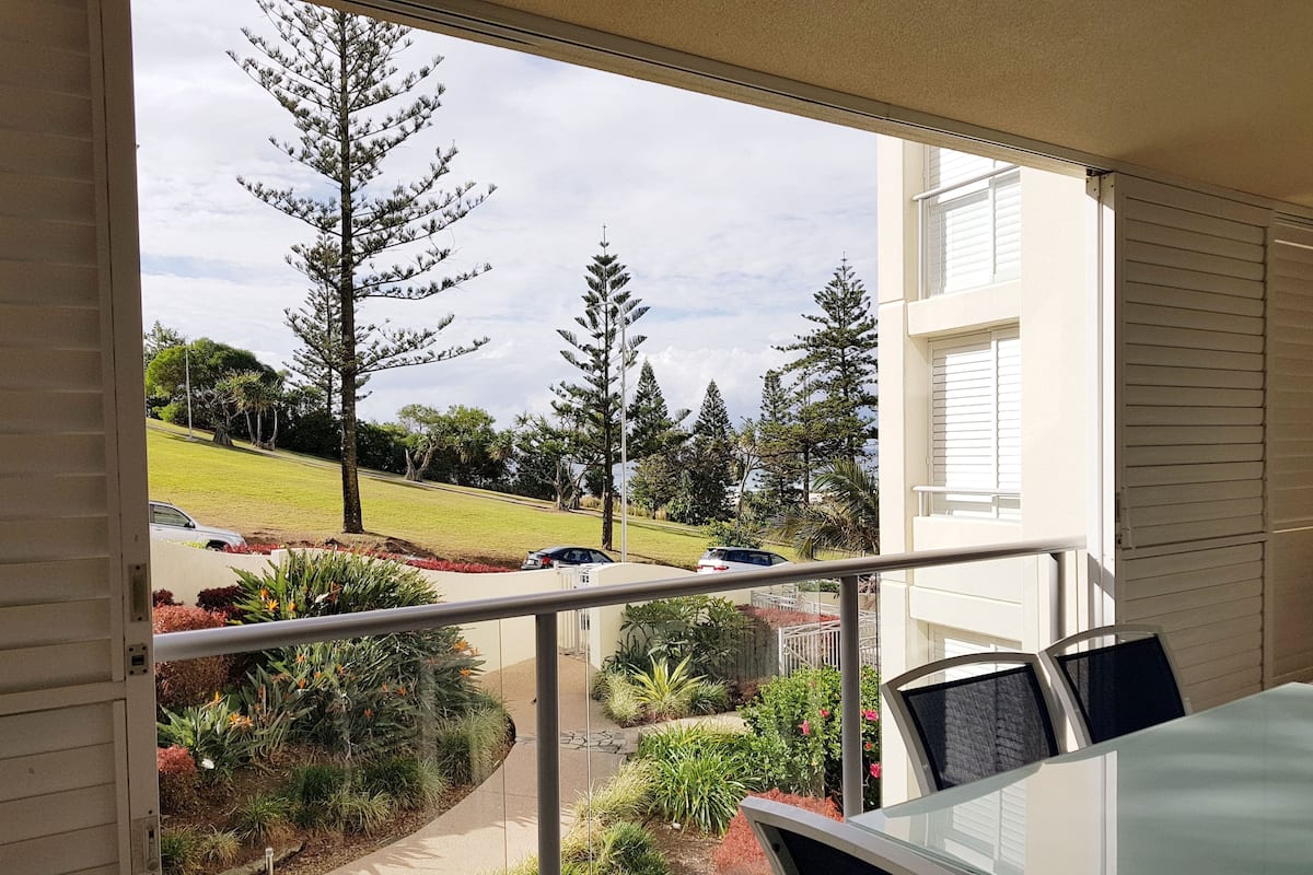 A balcony with a view of a landscaped garden, a car parked on the street, and a building with white siding.