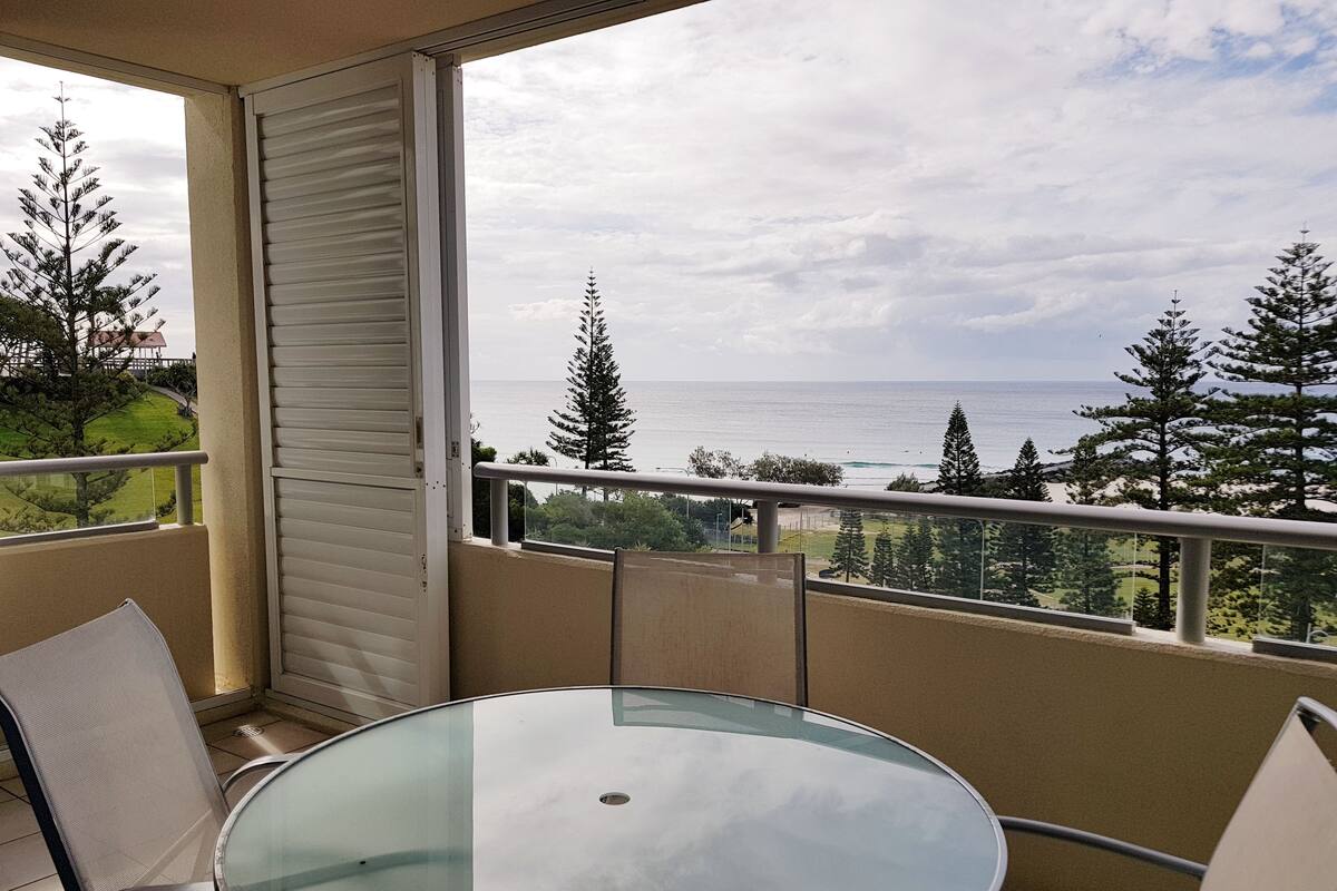 A balcony with a glass table and chairs, overlooking a coastal area with trees and the sea.