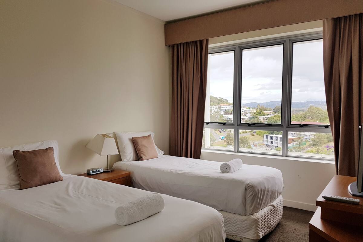 A hotel room with two beds, a nightstand with a lamp, a view of the city through a large window, and a wooden dresser.