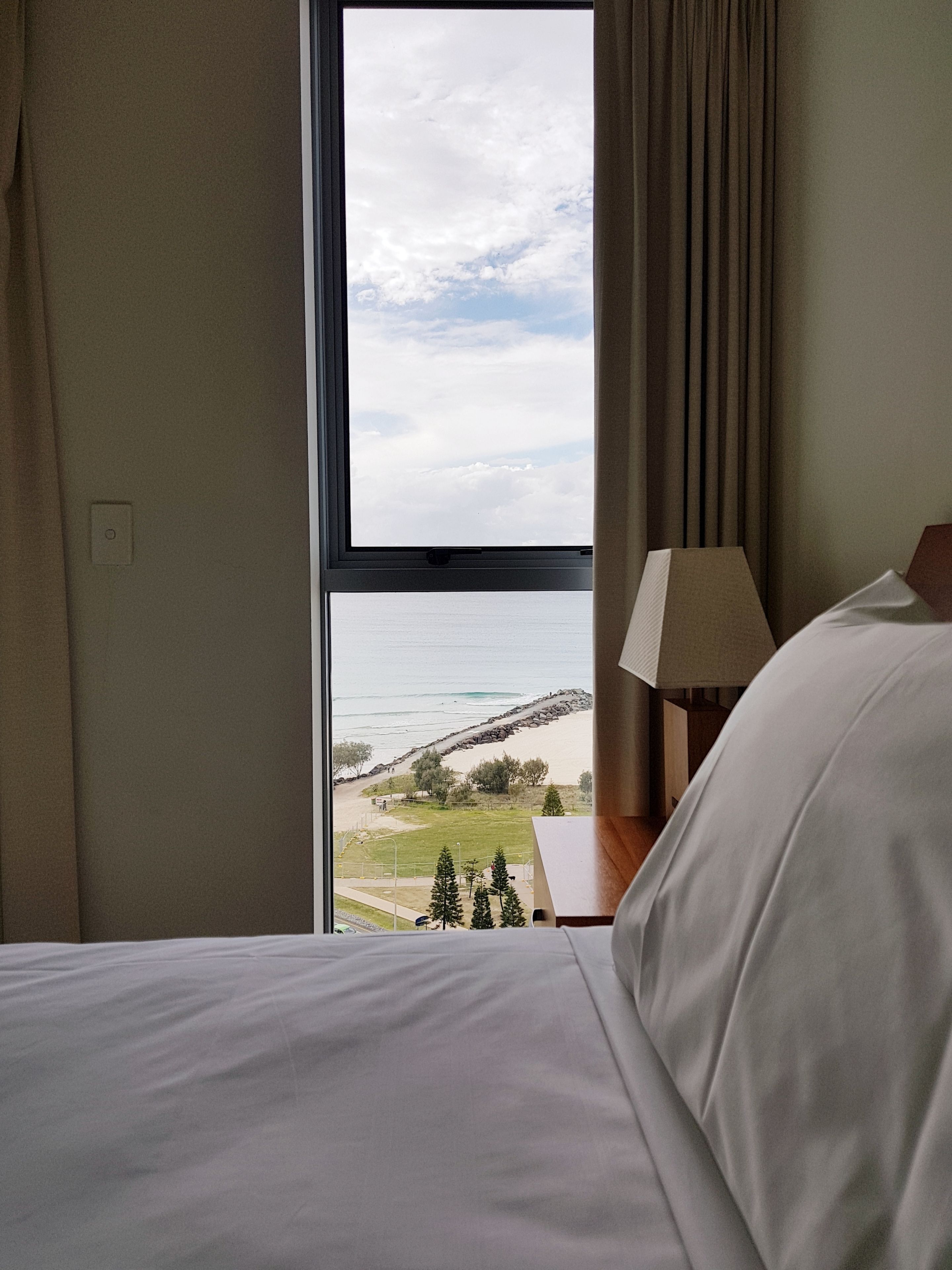 A hotel room with a bed, a nightstand with a lamp, and a view of the beach and ocean through a large window.