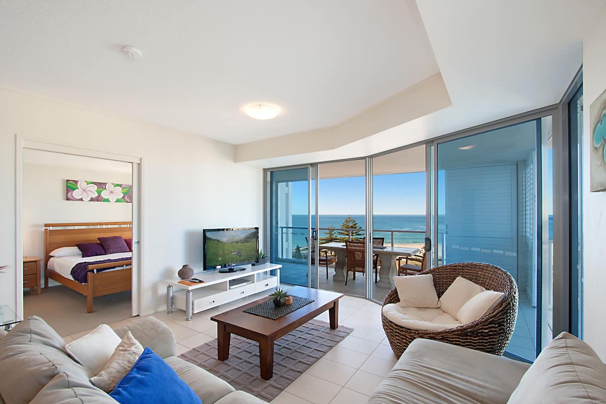 A modern living room with a bed, TV, and a balcony with a view of the ocean.