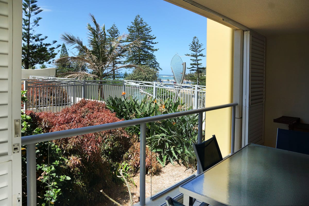 A balcony with a metal railing, a table, and chairs. There is a view of trees and a body of water in the background.