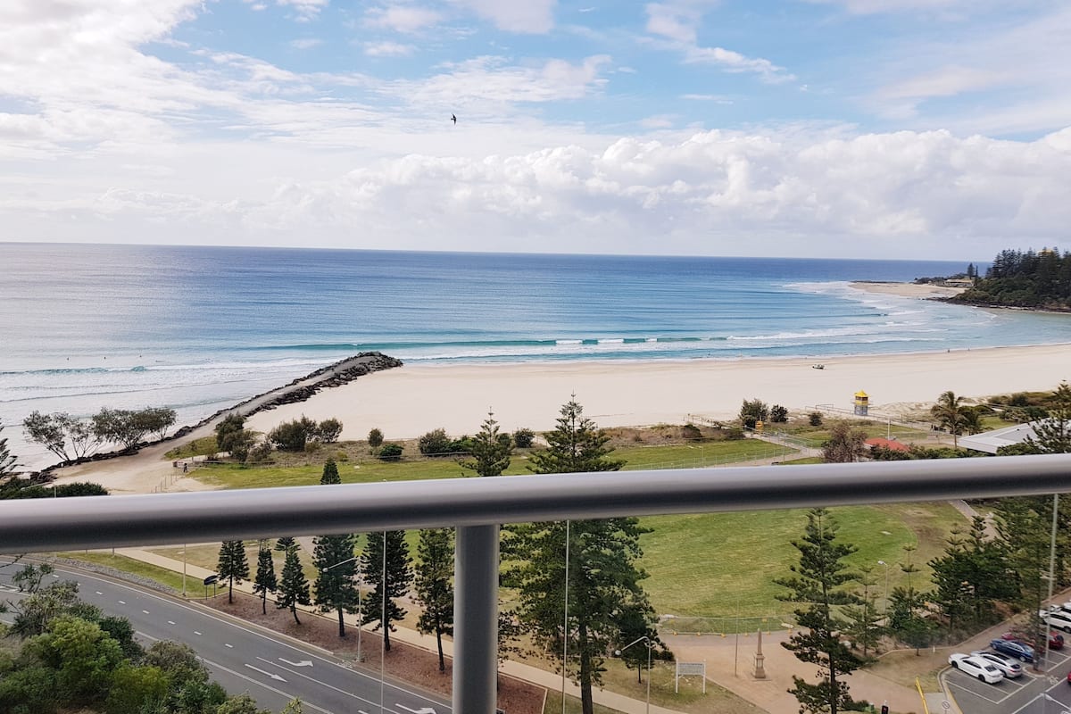 A coastal view from a balcony overlooking a beach, a road, and some greenery.