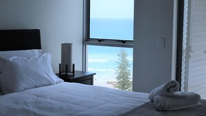 Une chambre à coucher avec un lit, une table de chevet, une lampe et une vue sur l’océan par une fenêtre ouverte.