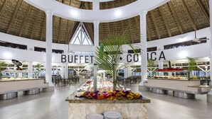 Een buffetgedeelte met verschillende food stations, een decoratieve plant in het midden en een zithoek met tafels en stoelen.