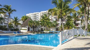 Una piscina con sillones de descanso, rodeada de palmeras y un edificio blanco con balcones.