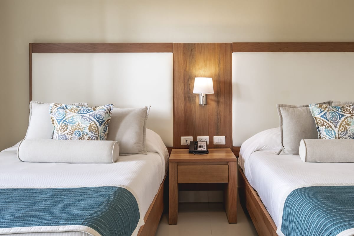 Een hotelkamer met twee bedden, elk voorzien van een gekleurd kussen en een blauwe plaid. Daartussen staat een houten nachtkastje met een lamp.
