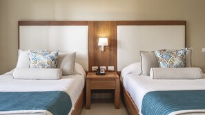 Habitación de hotel con dos camas, cada una con una almohada estampada y una manta azul. Hay una mesita de noche de madera con una lámpara entre las camas.
