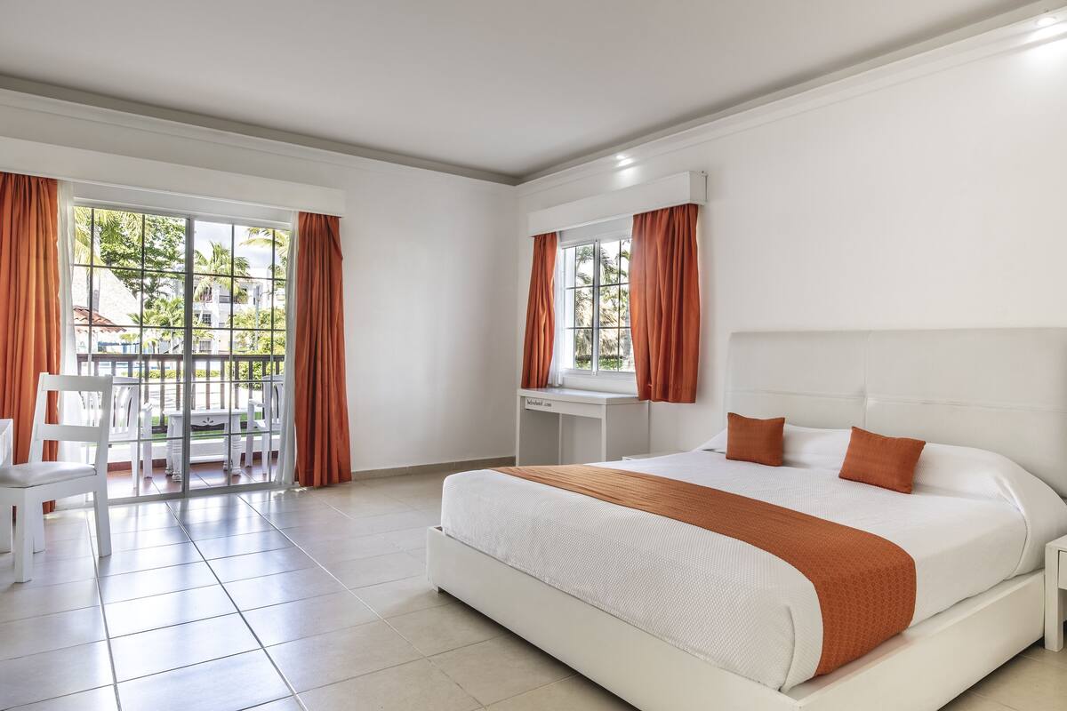 Een moderne hotelkamer met een groot bed, een bureau en een balkon met oranje gordijnen.