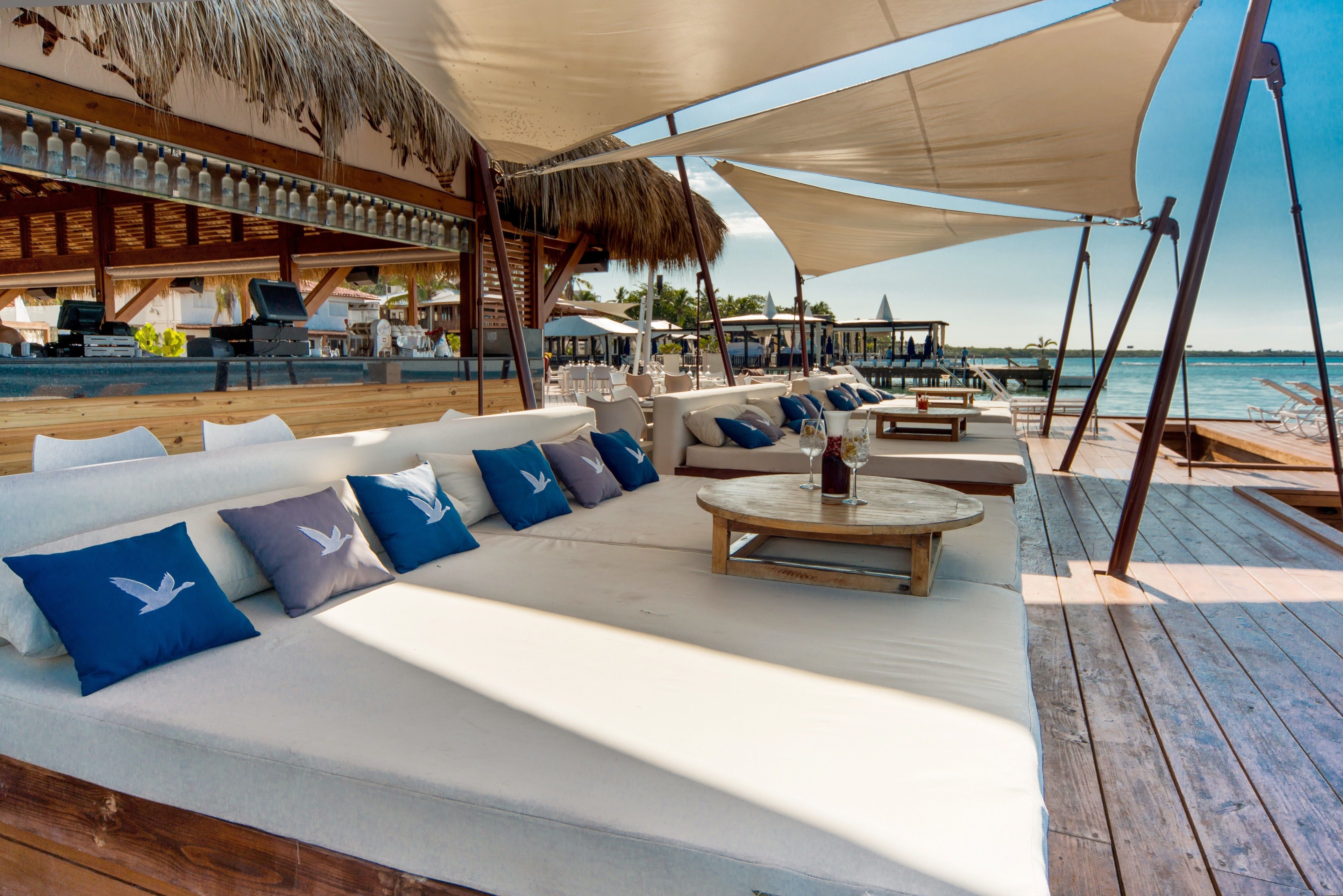 A beachside lounge with white cushioned seating, blue pillows, and a wooden table with a glass.