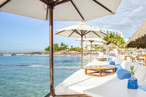 Un área de descanso frente a la playa, con asientos acolchados blancos y mesas de madera, con vista a un mar azul claro y una playa de arena con palmeras.