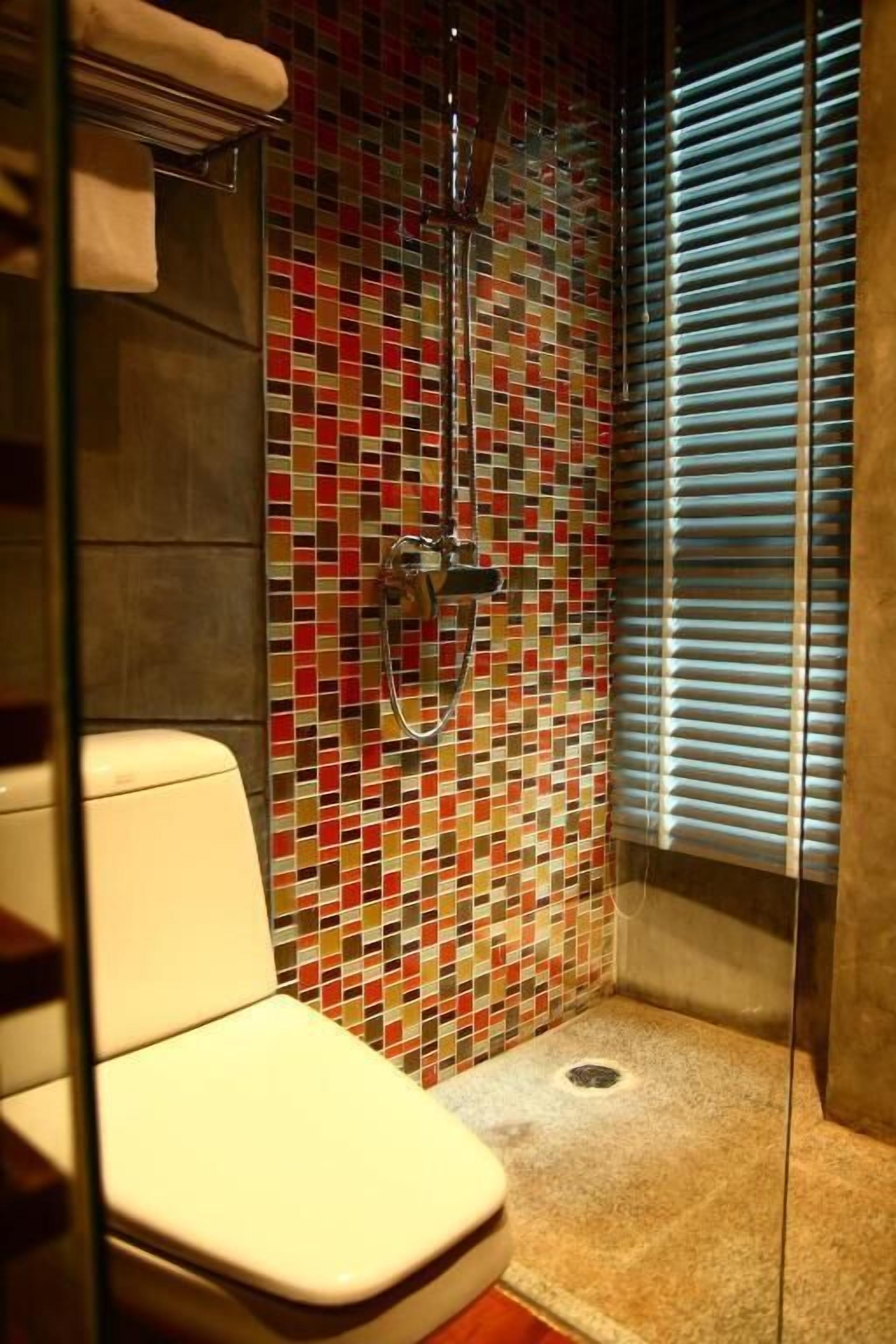 A small bathroom with a tiled shower wall, a toilet, and a window with blinds.