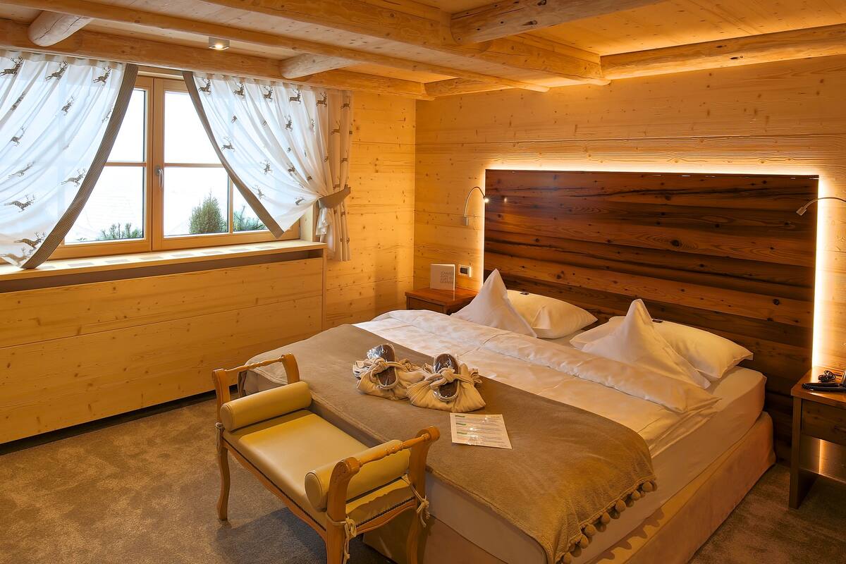 A wooden cabin bedroom with a bed, a chair, a bedside table with a lamp, and a window with curtains.