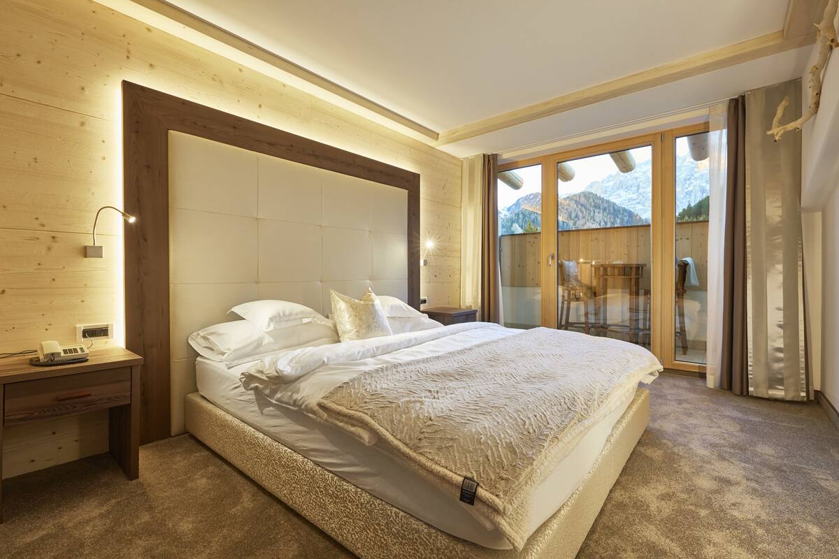 A bedroom with a large bed, wooden headboard, bedside tables, and a view of mountains through a glass door.