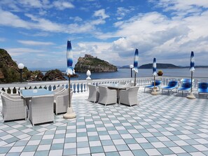 A terrace with a checkered floor, wicker furniture, and umbrellas. A castle is visible in the background. - Hotel Da Maria (Ischia)