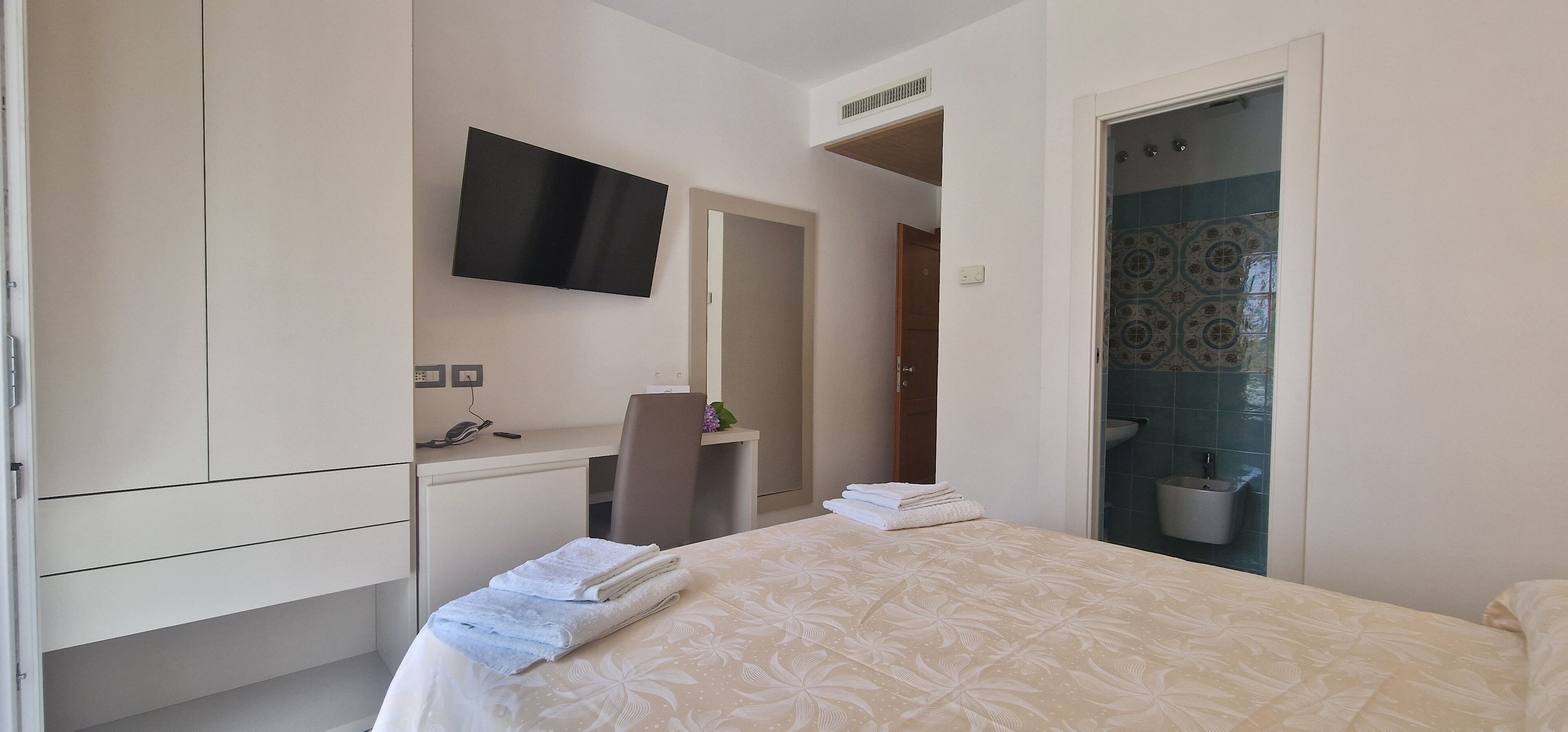 A hotel room with a bed, a desk with a chair, a TV mounted on the wall, and a bathroom with a shower and sink.
