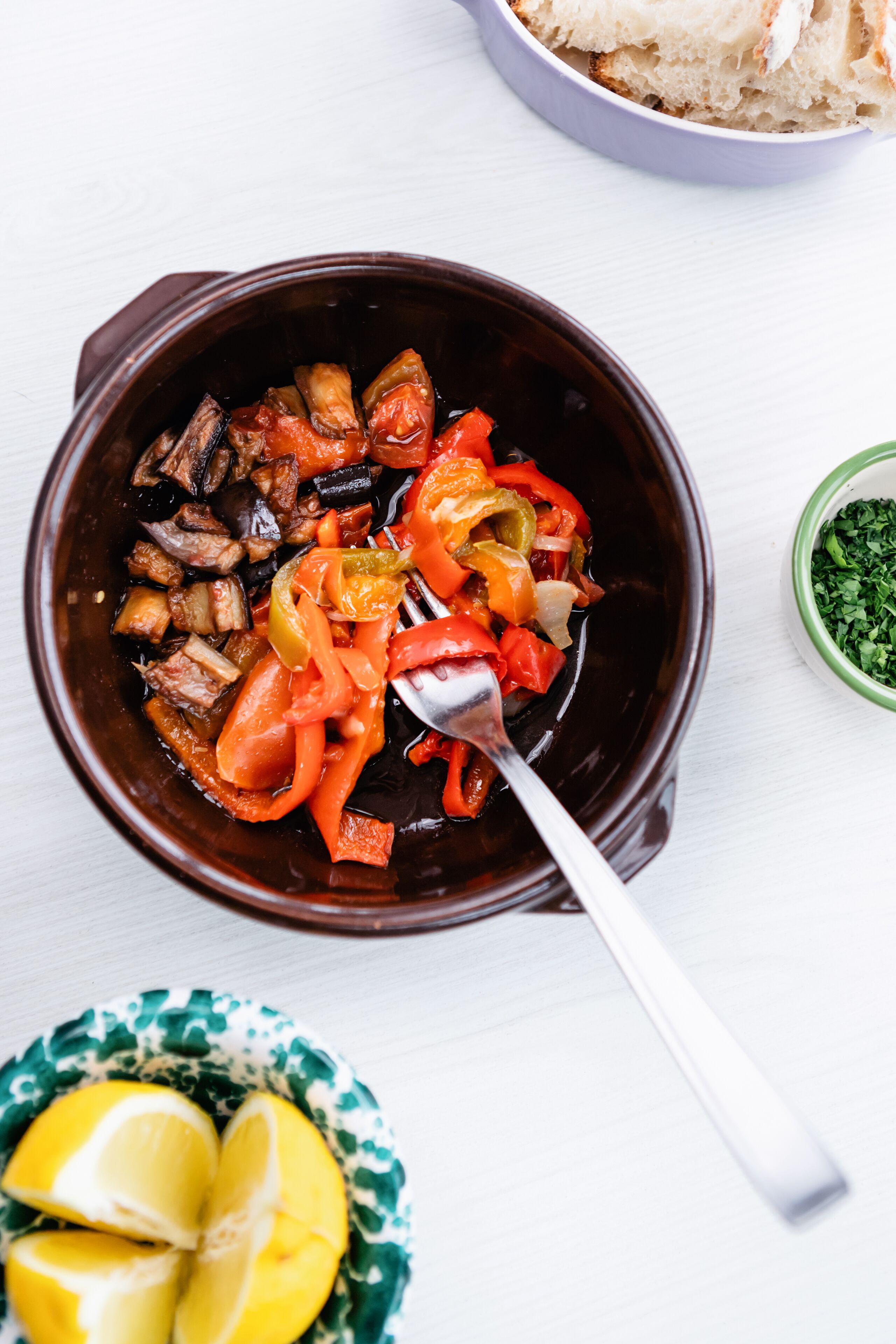 A bowl of mixed vegetables with a fork, a bowl of bread, and a small bowl of herbs.