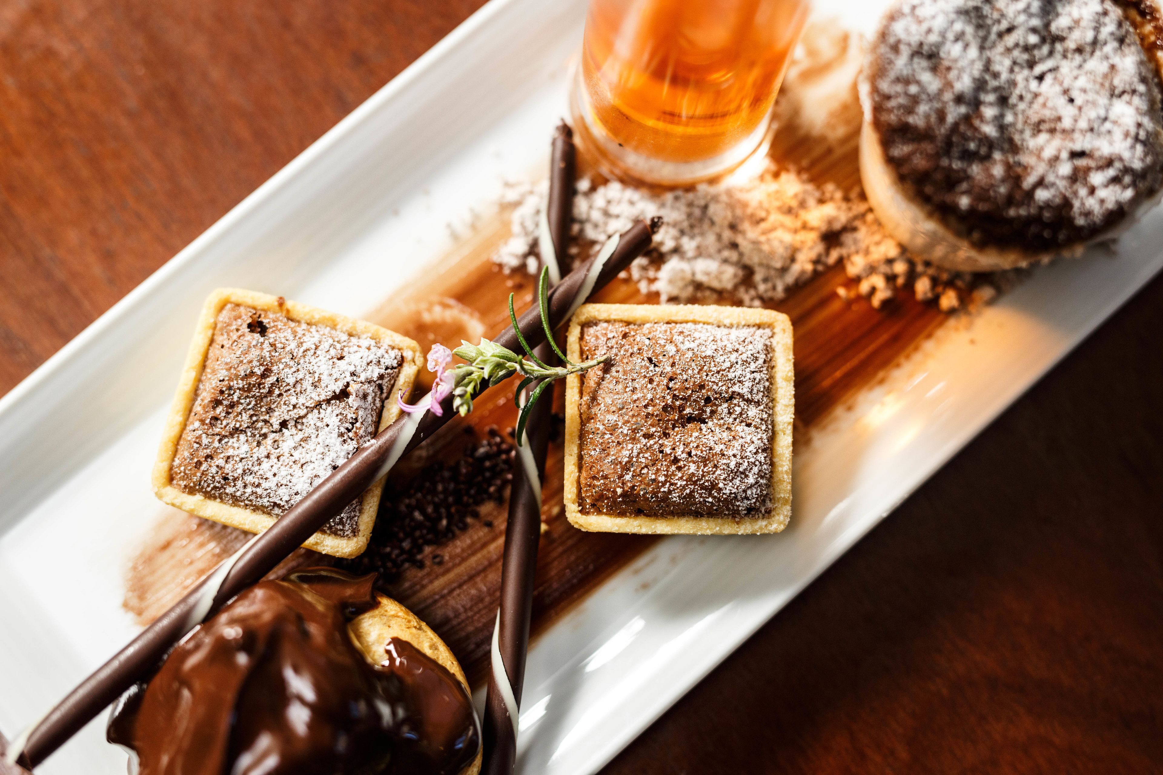 Un plato de postres con dos pastelitos cuadrados espolvoreados con azúcar glas, un dulce cubierto de chocolate y un vaso de bebida color ámbar.