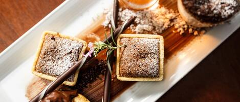Un plato de postres con dos pastelitos cuadrados espolvoreados con azúcar glas, un dulce cubierto de chocolate y un vaso de bebida color ámbar.