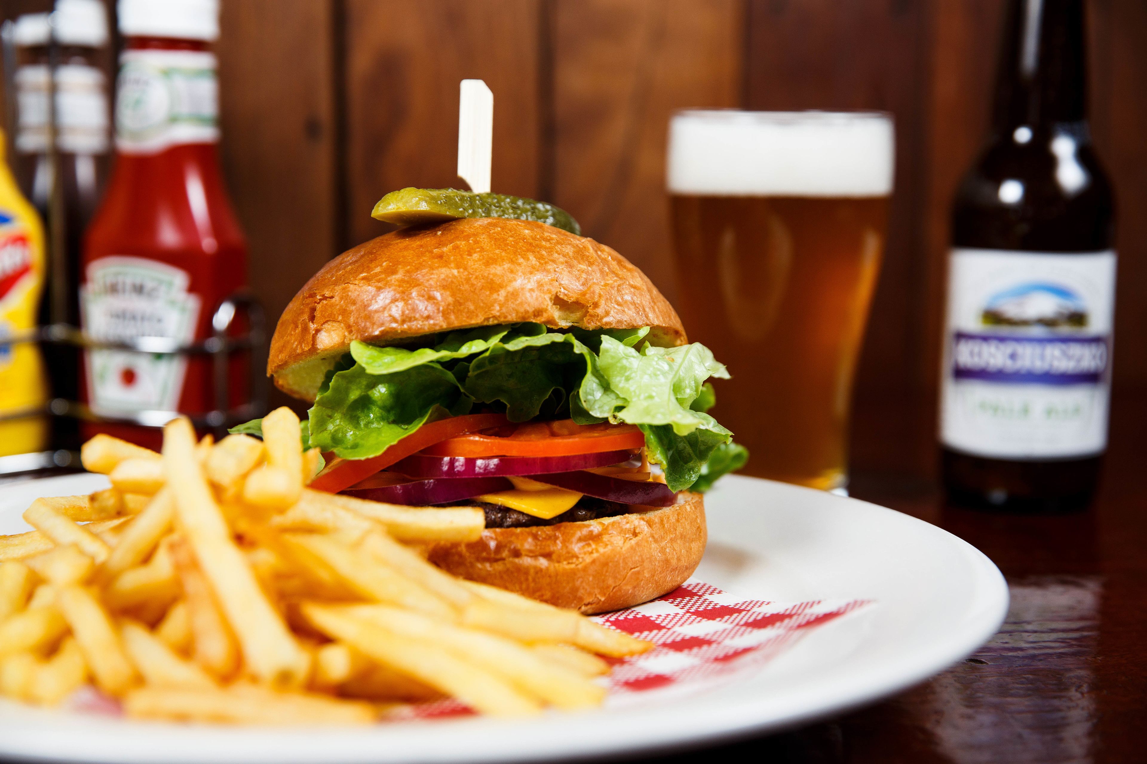 A burger with fries on a plate, a glass of beer, and condiments in the background.