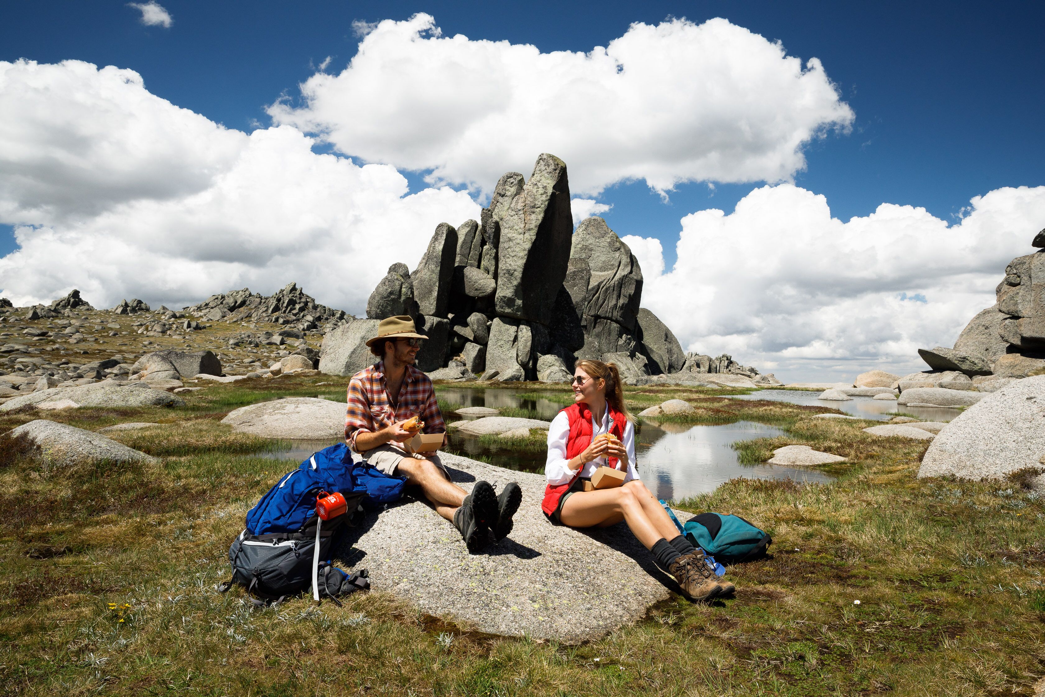 Two people sitting on rocks with a mountainous landscape and a body of water in the background.
