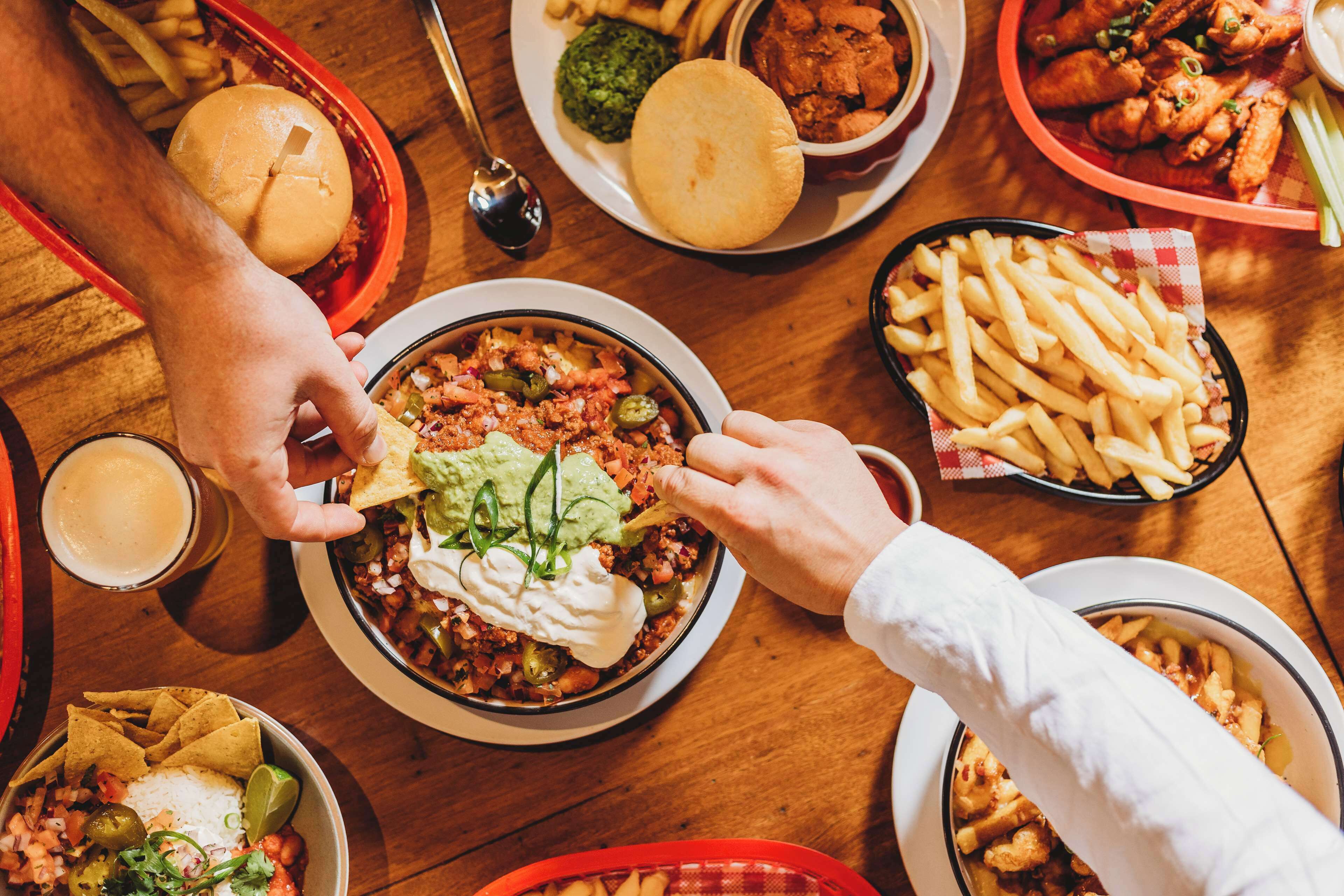Una mesa con varios platos, incluyendo nachos, una hamburguesa y un tazón de chili cubierto de guacamole y crema agria.