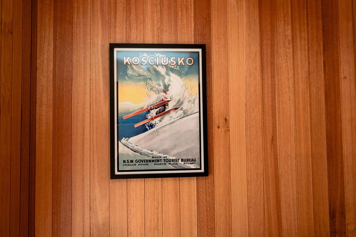 A framed poster of a ski scene with the text "KOSCIUSKO" and "BOOK AT N.S.W. GOVERNMENT TOURIST BUREAU" on a wooden wall.