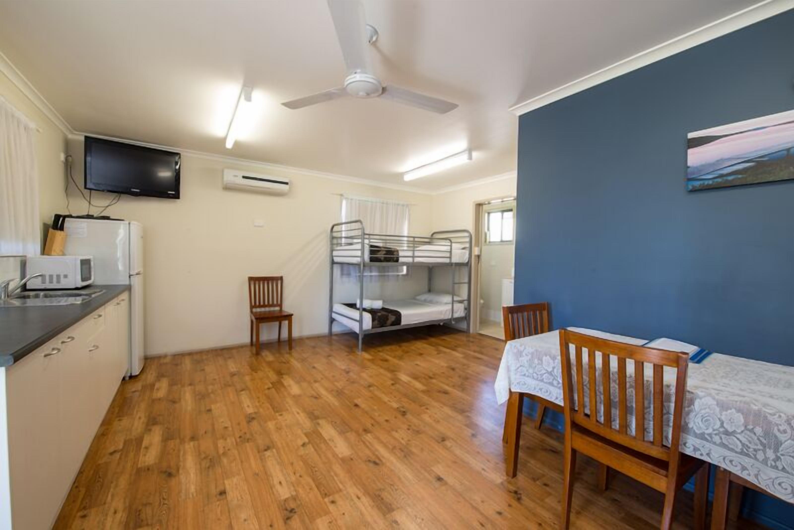 A compact room with a bunk bed, a dining table with chairs, a television mounted on the wall, and a ceiling fan.