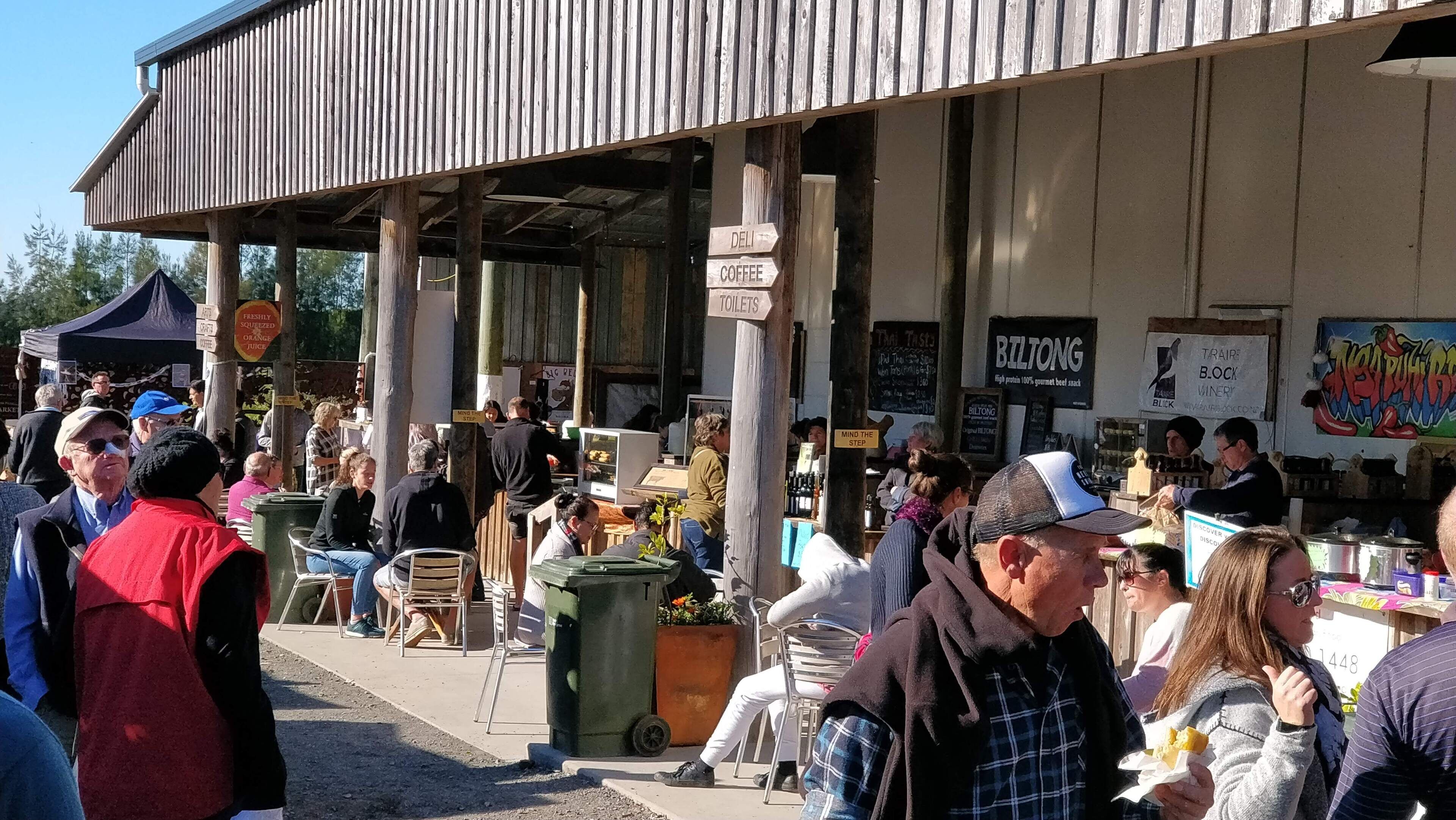 A covered outdoor seating area with wooden pillars, a sign indicating a deli, coffee, and toilets, and a mural on the wall.