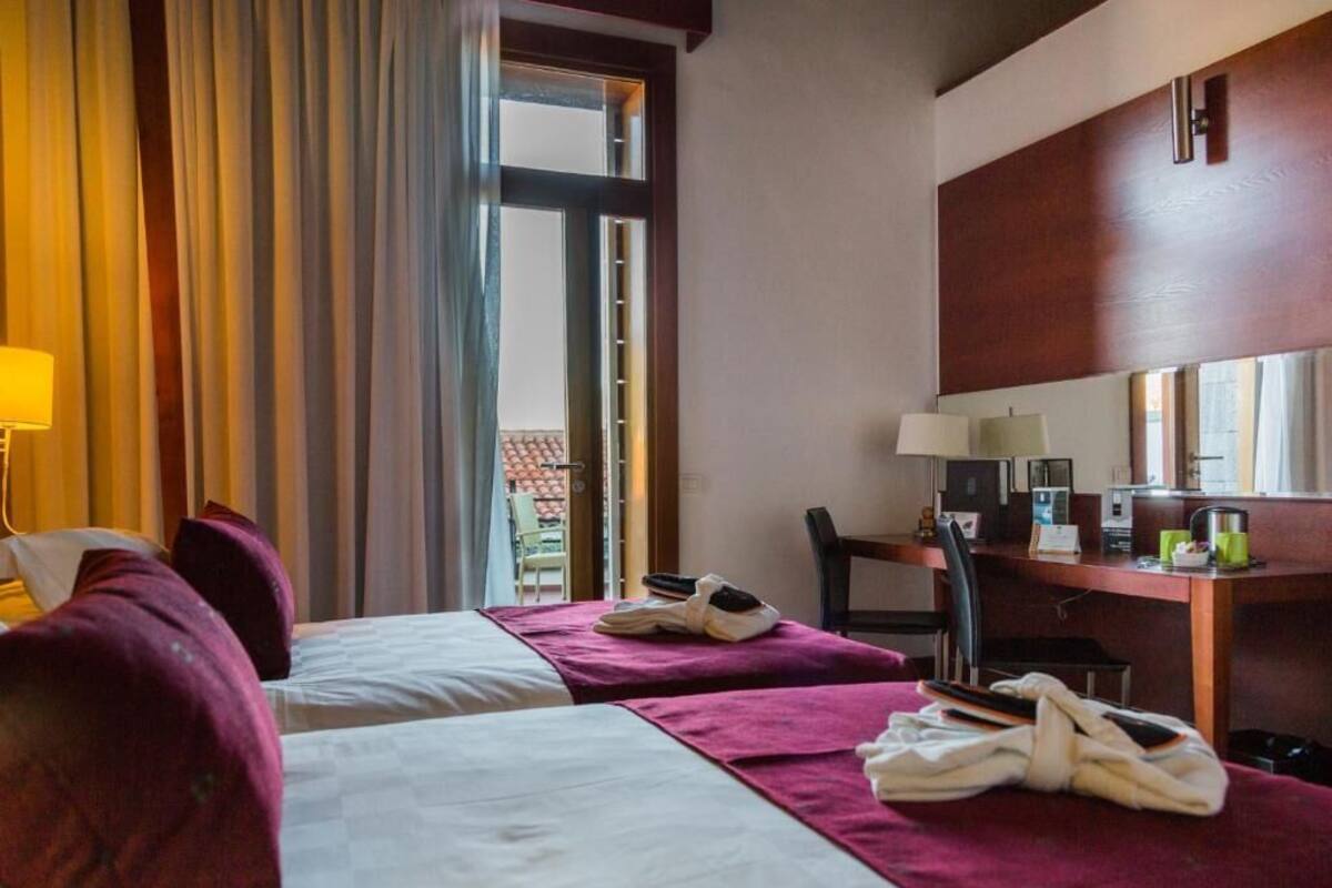 A hotel room with two beds, a desk, a chair, a lamp, and a window with curtains.