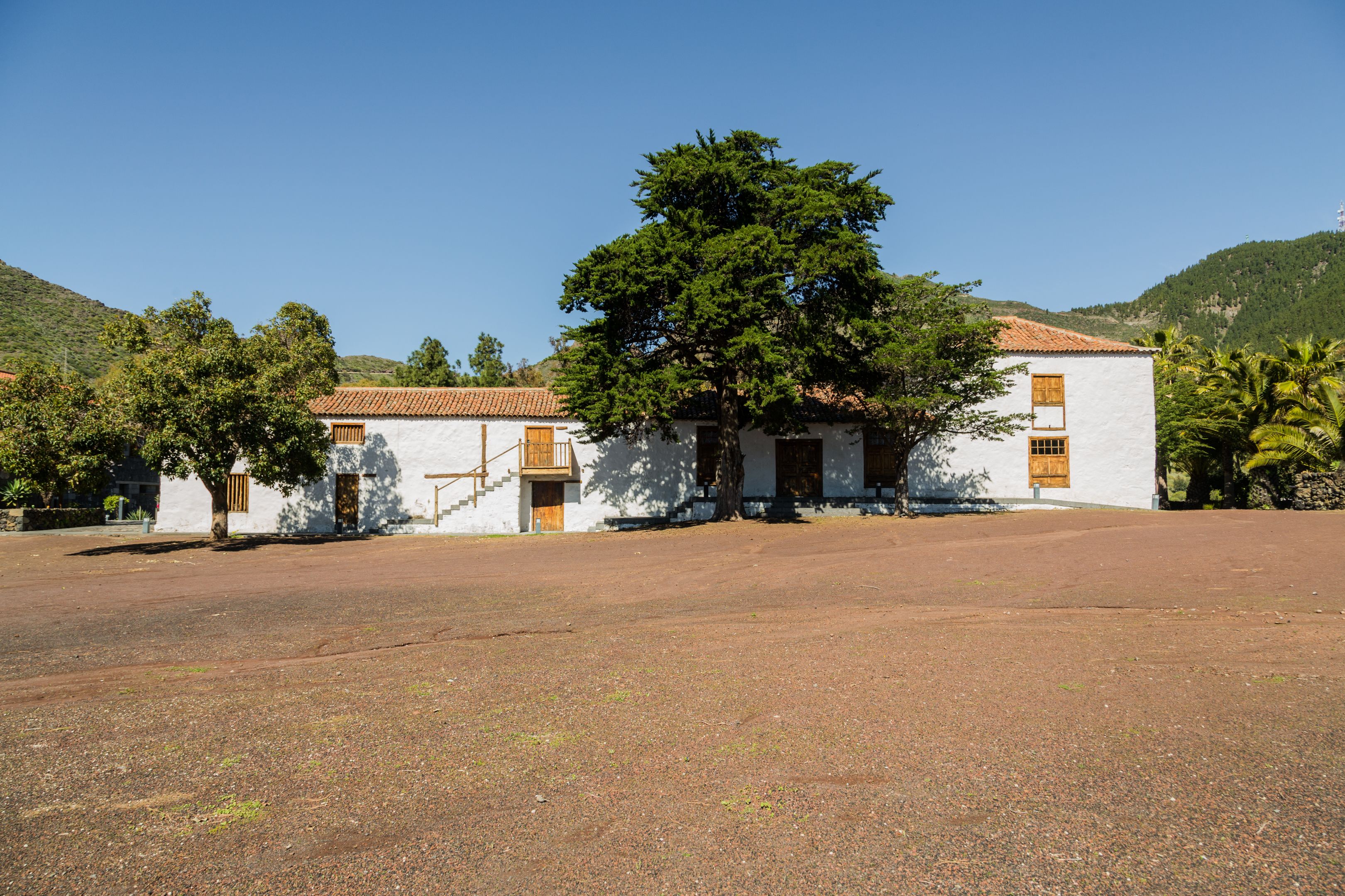 A building with a white facade and red-tiled roof, surrounded by trees and a hilly landscape.
