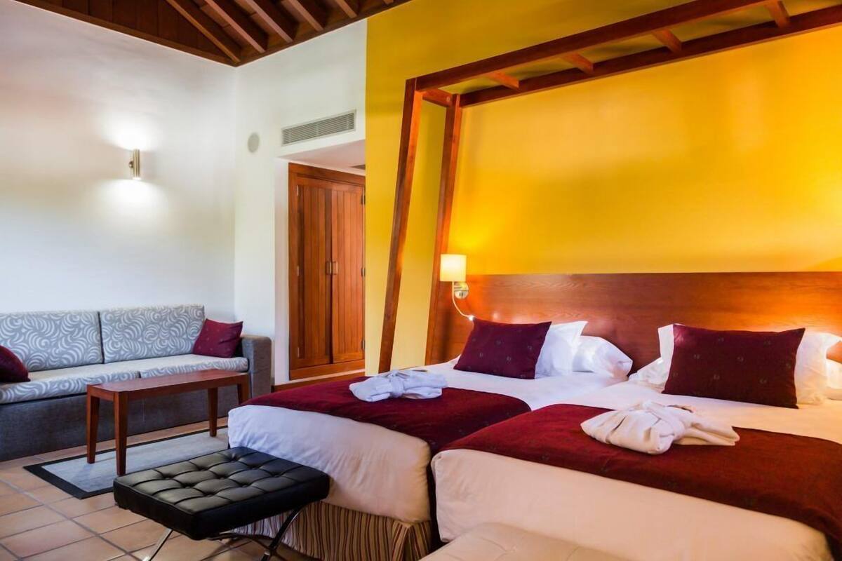 A hotel room with a sofa, two beds, a wooden headboard, and a nightstand.
