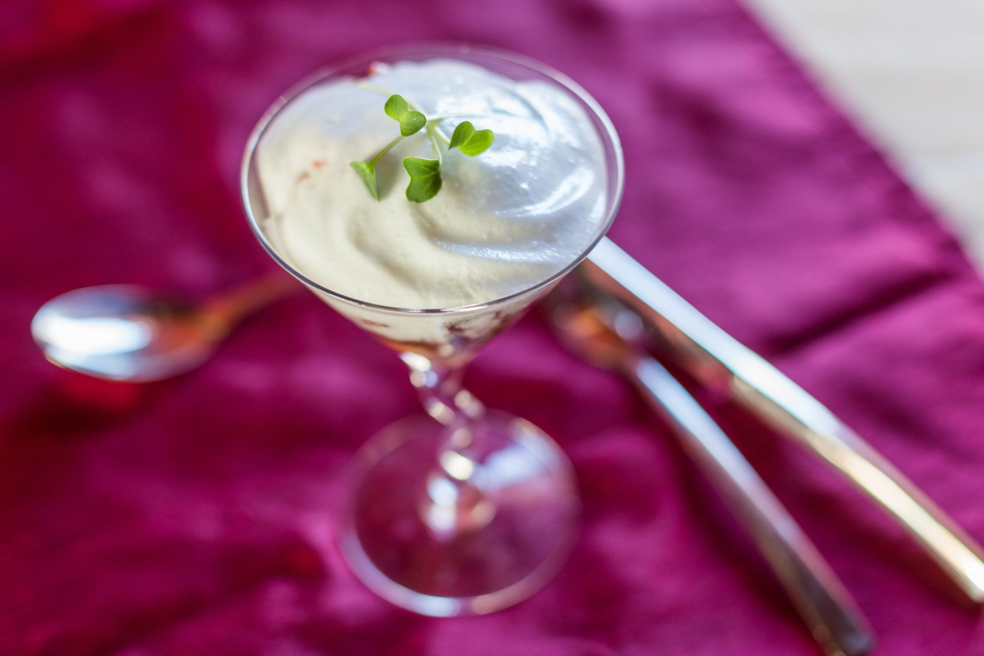 Una copa de martini con una bebida blanca cremosa, adornada con una pequeña hoja verde, sobre un paño de color morado intenso.
