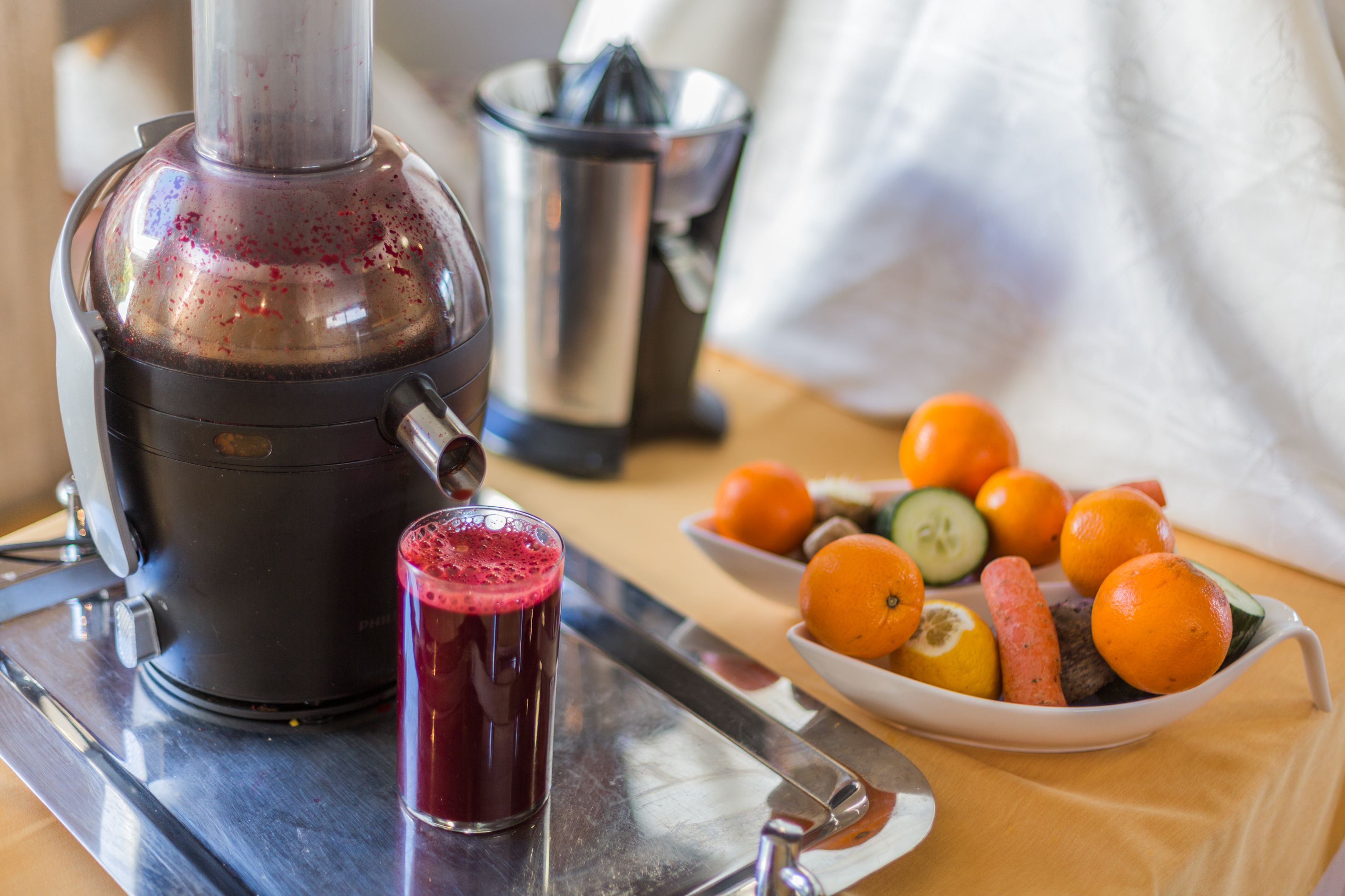 A juicer with a glass of juice, a bowl of fruits, and a coffee maker on a kitchen counter.