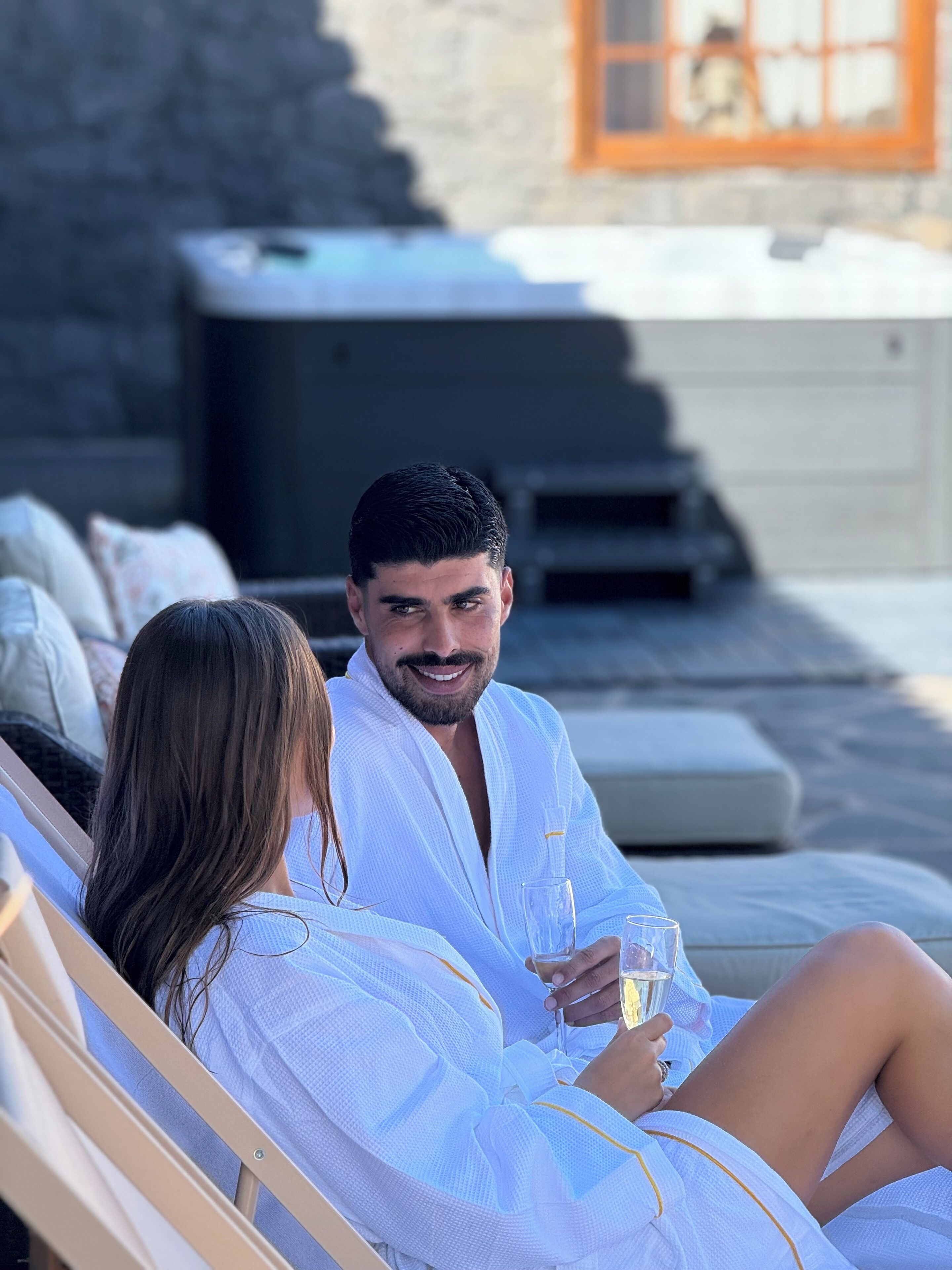 Un couple se détend sur des chaises longues au soleil, un verre à la main, avec un mur de pierres et un jacuzzi en arrière-plan.