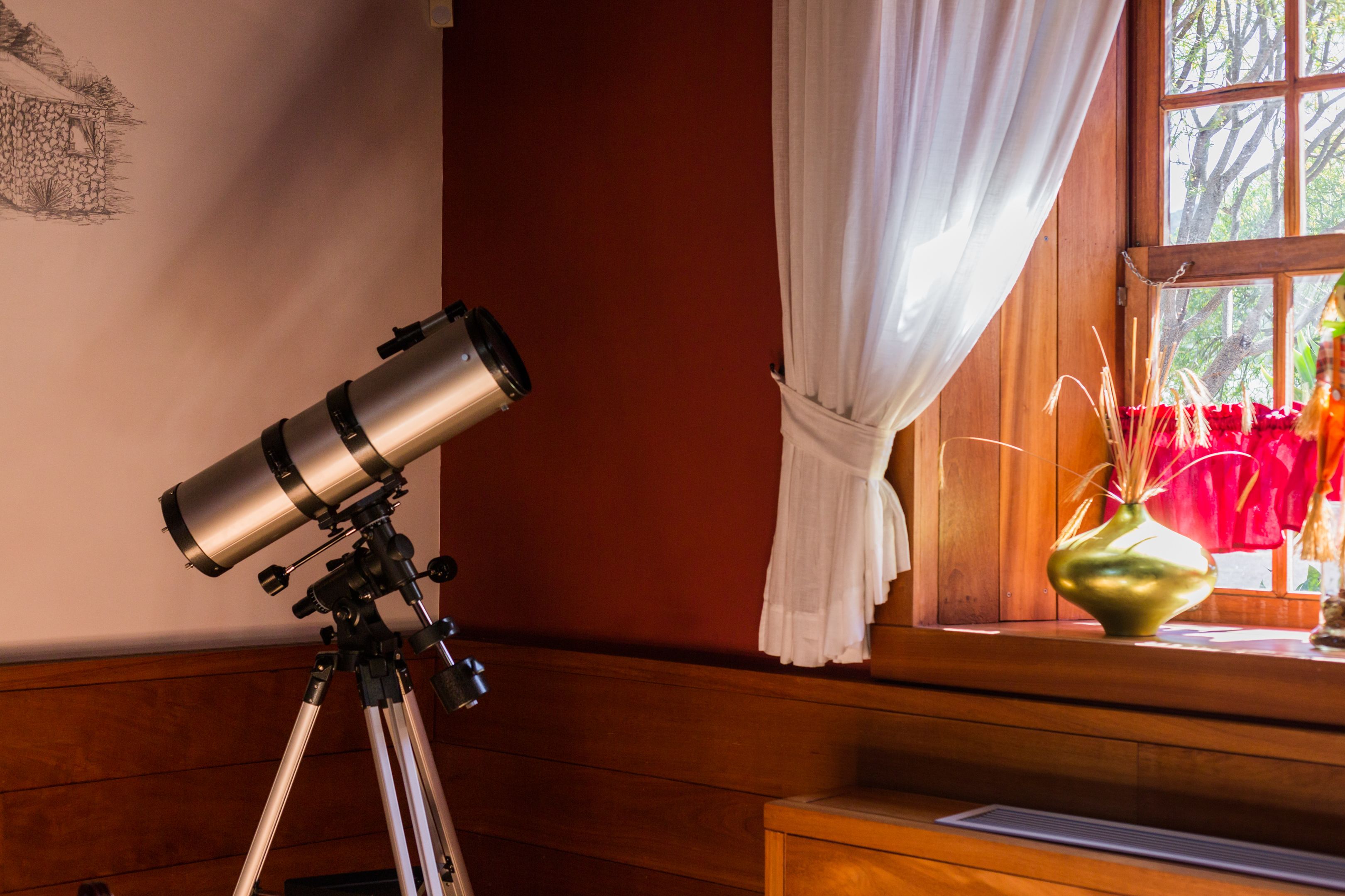 A telescope on a tripod in a room with wooden walls and a window with curtains.