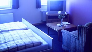 Quarto de hotel com uma cama, uma cadeira, uma mesinha com um vaso de flores, uma televisão e uma janela com cortinas.