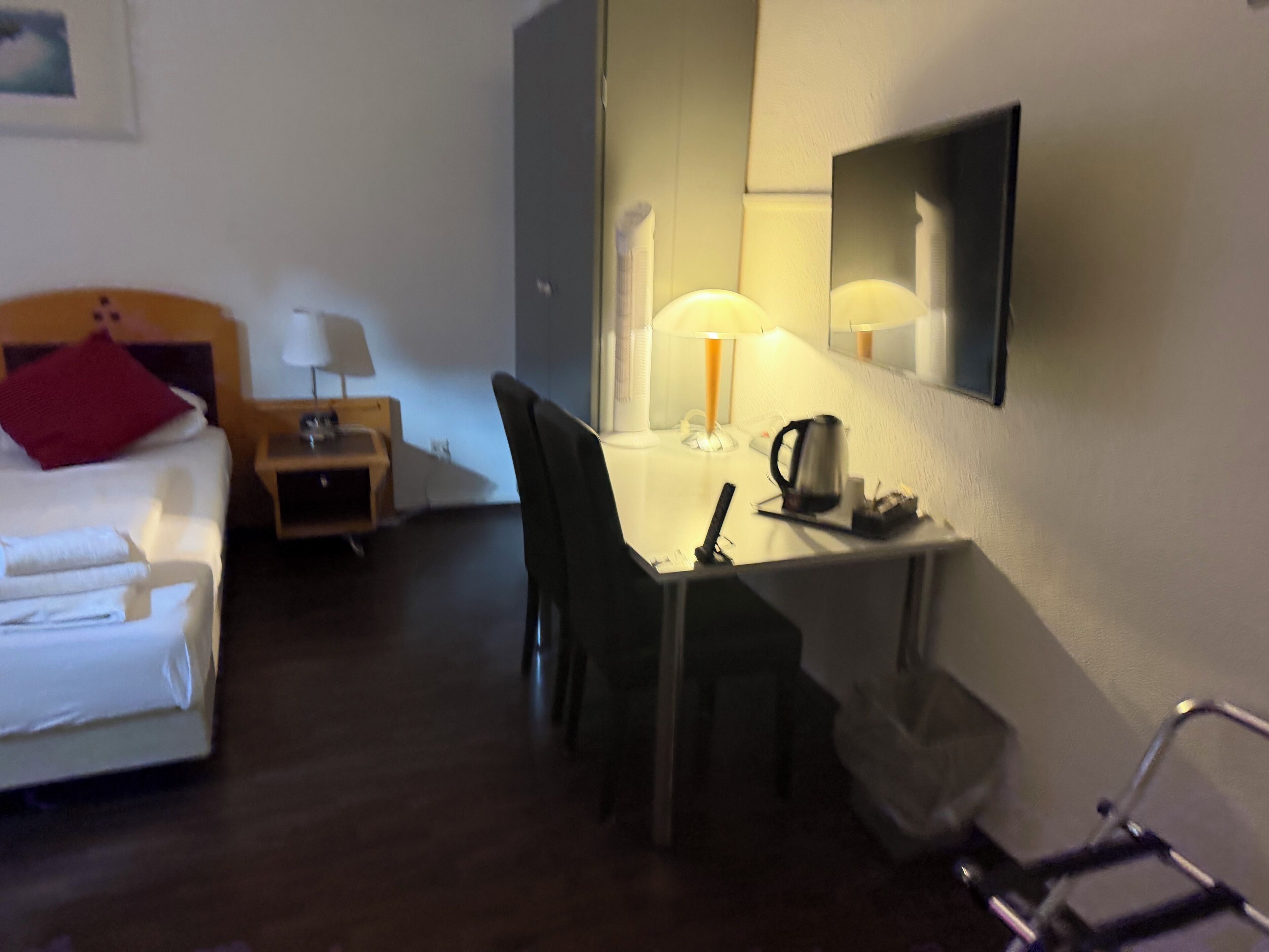 A hotel room with a bed, a desk, a chair, a lamp, a kettle, and a mirror.