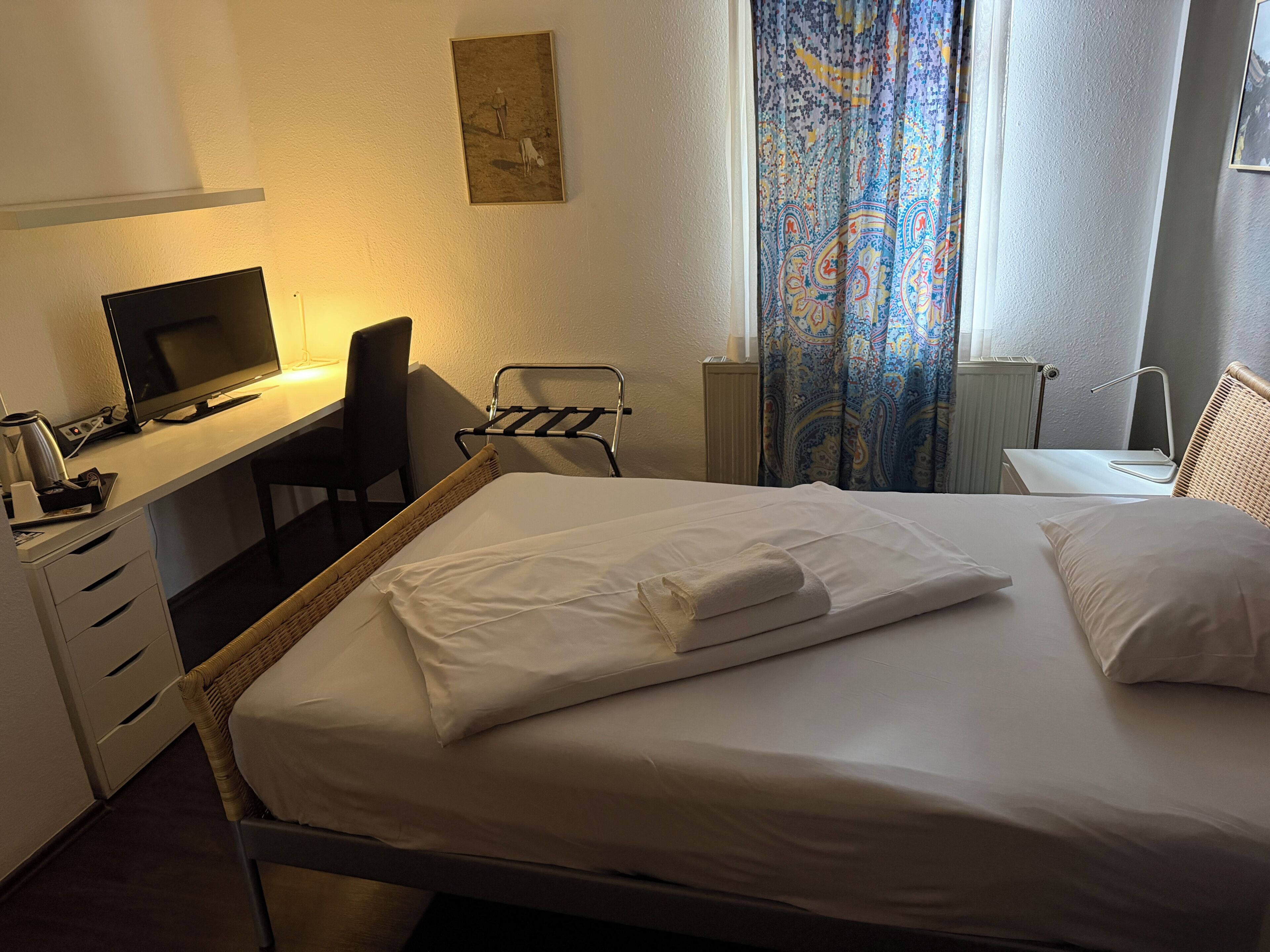 A hotel room with a bed, a desk with a computer, a chair, a radiator, and a window with curtains.
